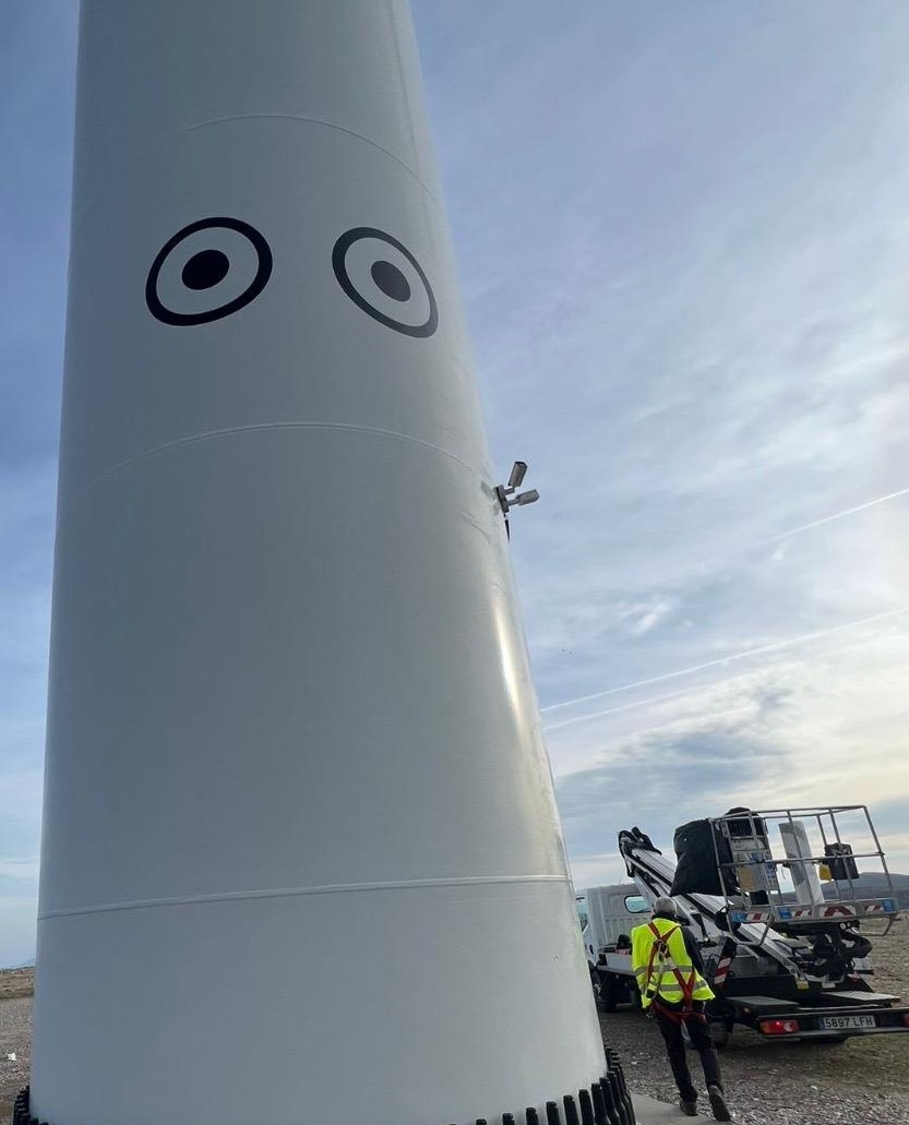 Vinilos con forma de ojos en una torre de aerogenerador, con personal y plataforma de trabajo al pie